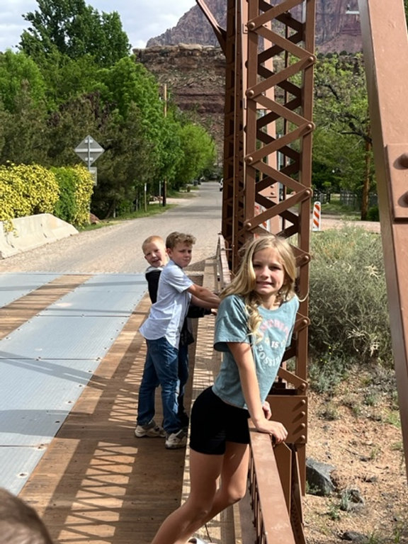 Two children standing on a bridge in the desert, surrounded by vast sandy dunes and a clear blue sky above. Version 1: Two kids on a desert bridge, surrounded by sandy dunes and a clear blue sky. Version 2: A bridge in the desert with two children standing on it, surrounded by sandy dunes and a clear blue sky. Version 3: Two children standing on a bridge in the desert, with sandy dunes and a clear blue sky in the background.